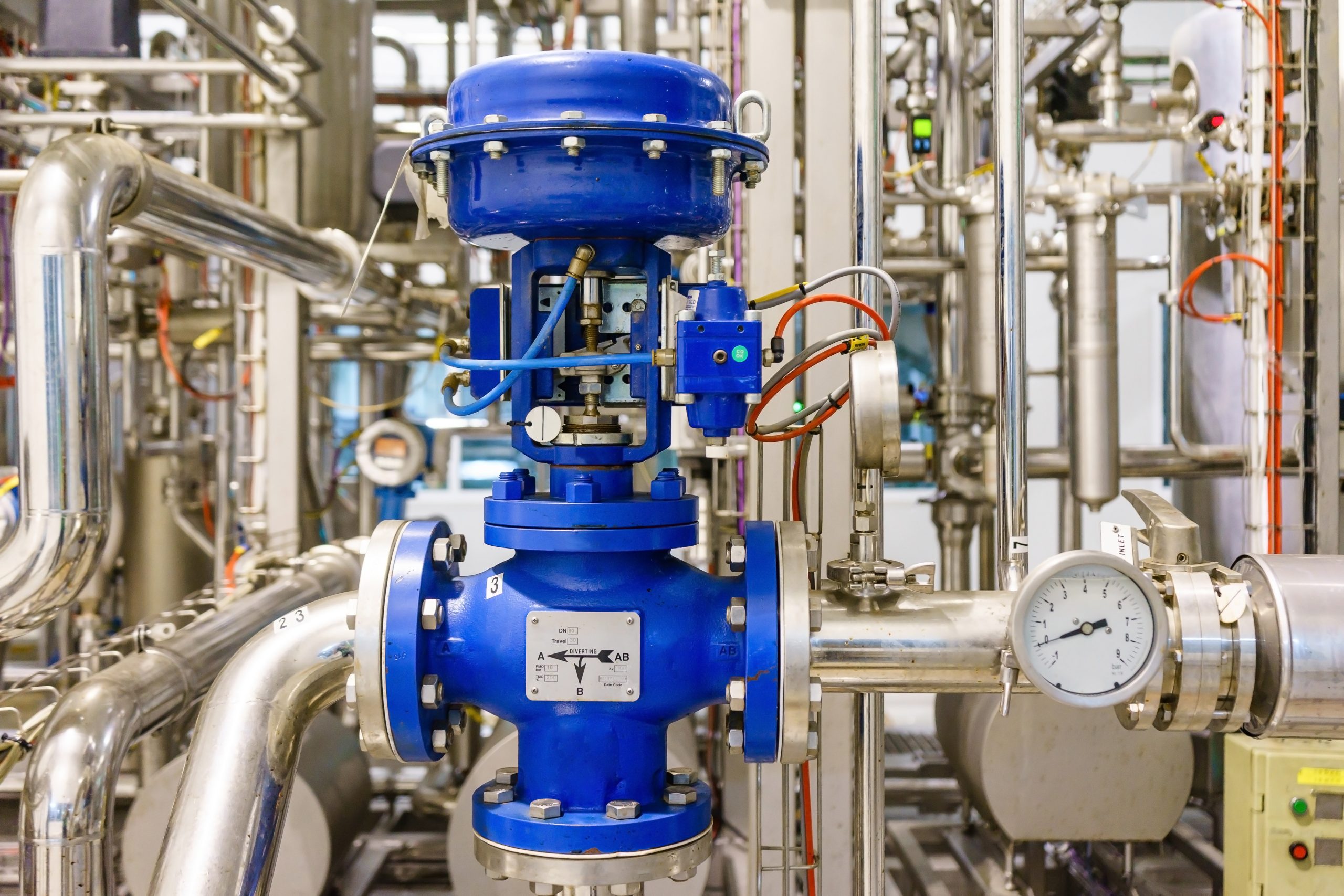 Blue pneumatic control valve installed in a stainless steel steam pipe system with pressure gauge and industrial piping.