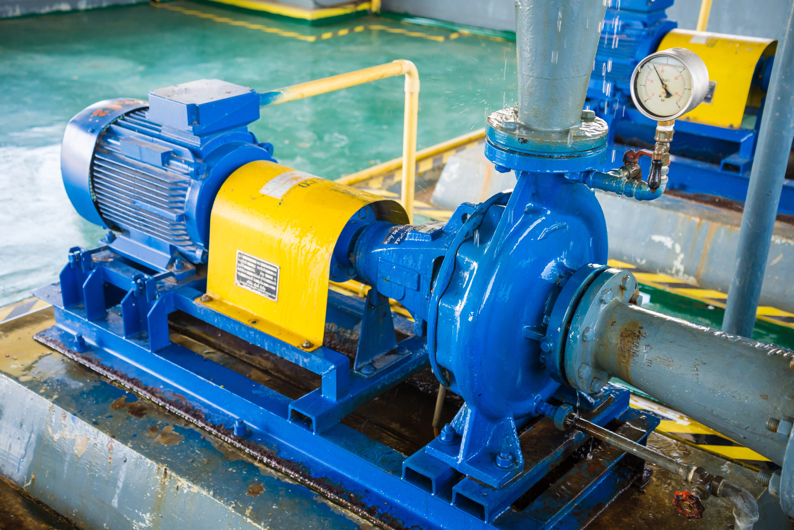 Blue industrial centrifugal pump with attached electric motor and pressure gauge connected to metal piping in a mechanical facility.