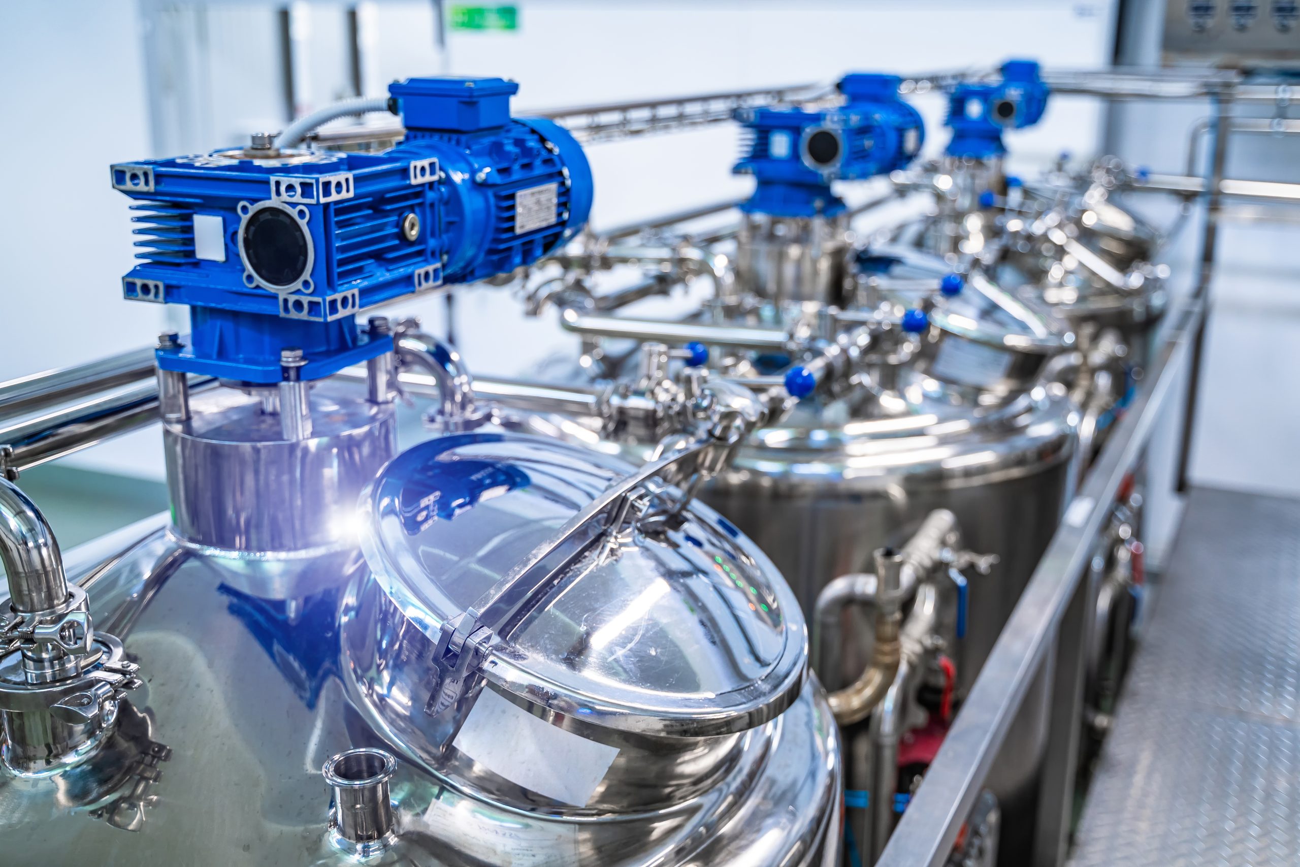 Stainless steel industrial mixing tanks with blue motorized agitators inside a clean manufacturing facility.