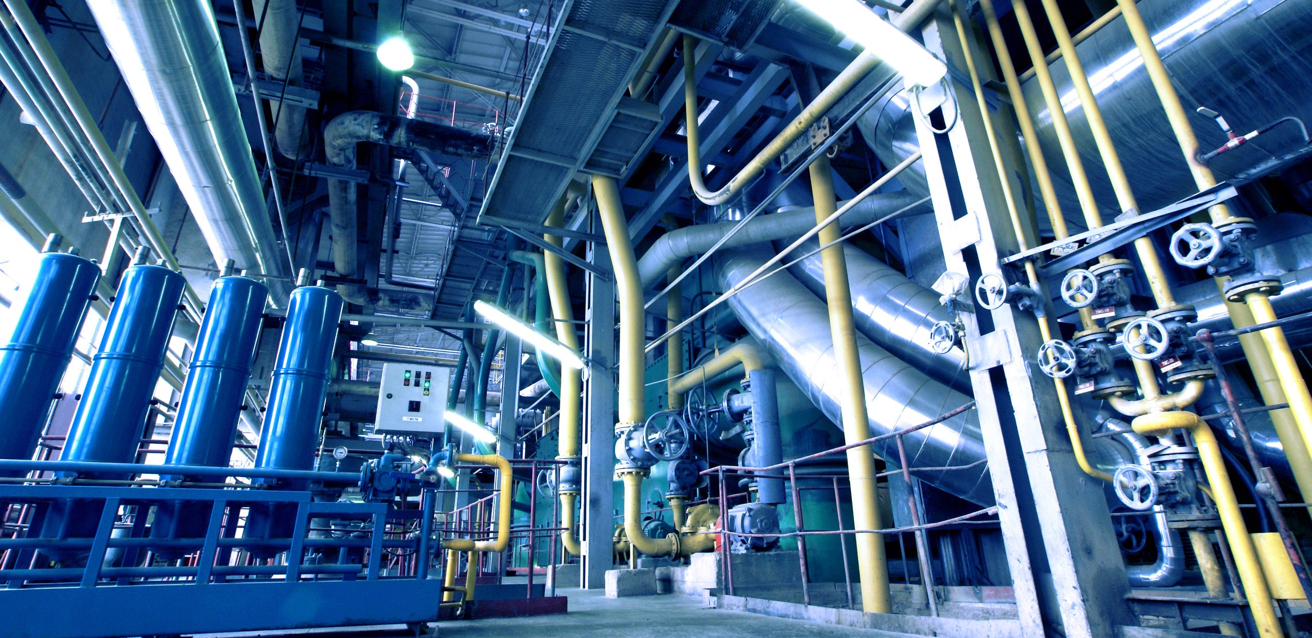 Interior of a modern industrial power plant with blue equipment, yellow support structures, pipes, cables, and control valves.