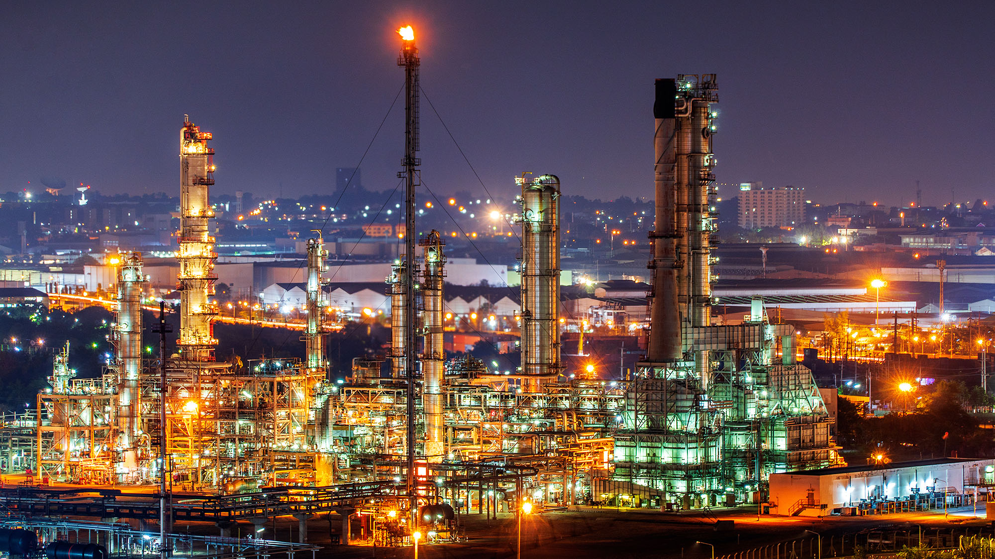 Night view of an illuminated industrial refinery with tall towers, pipes, and a burning flare stack.