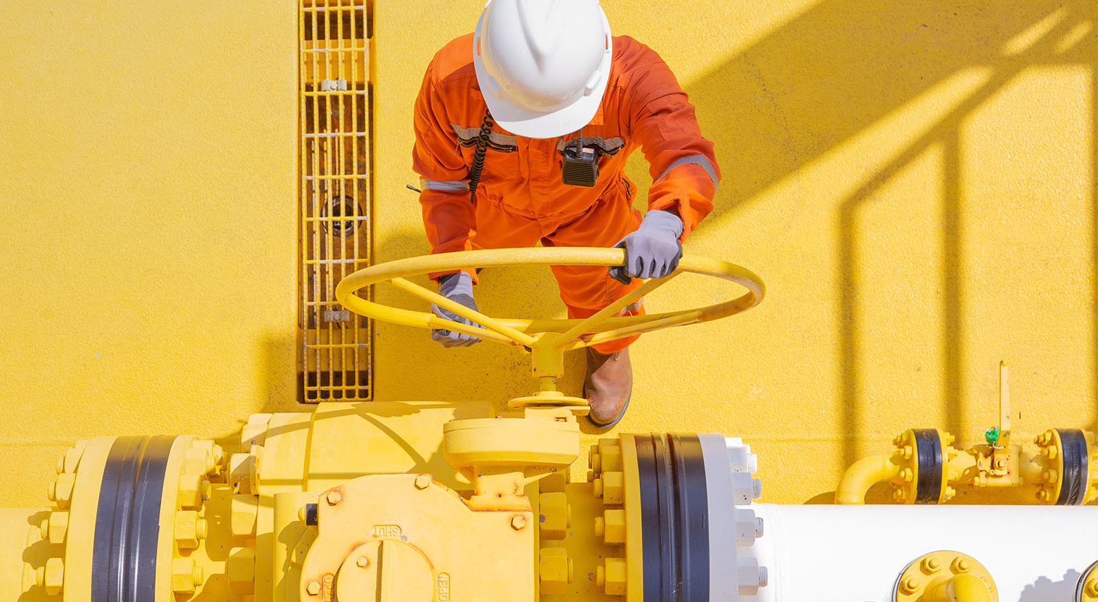 Worker in orange safety gear and hard hat turning a large yellow valve on an industrial pipeline, viewed from above.