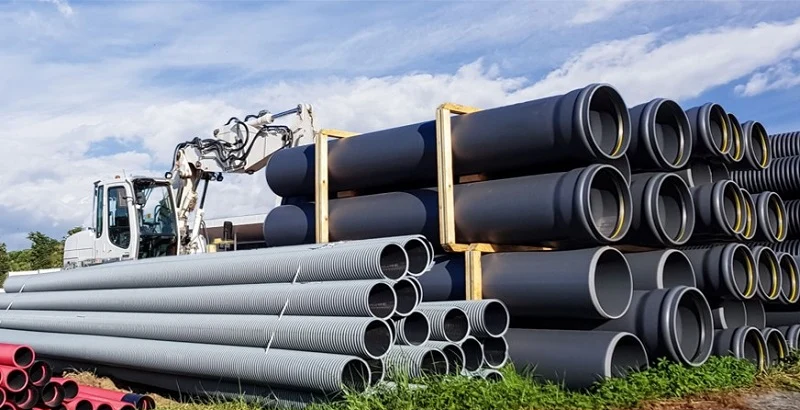 Stacks of large‑diameter non‑metallic pipes stored outdoors at an industrial site with heavy equipment in the background.